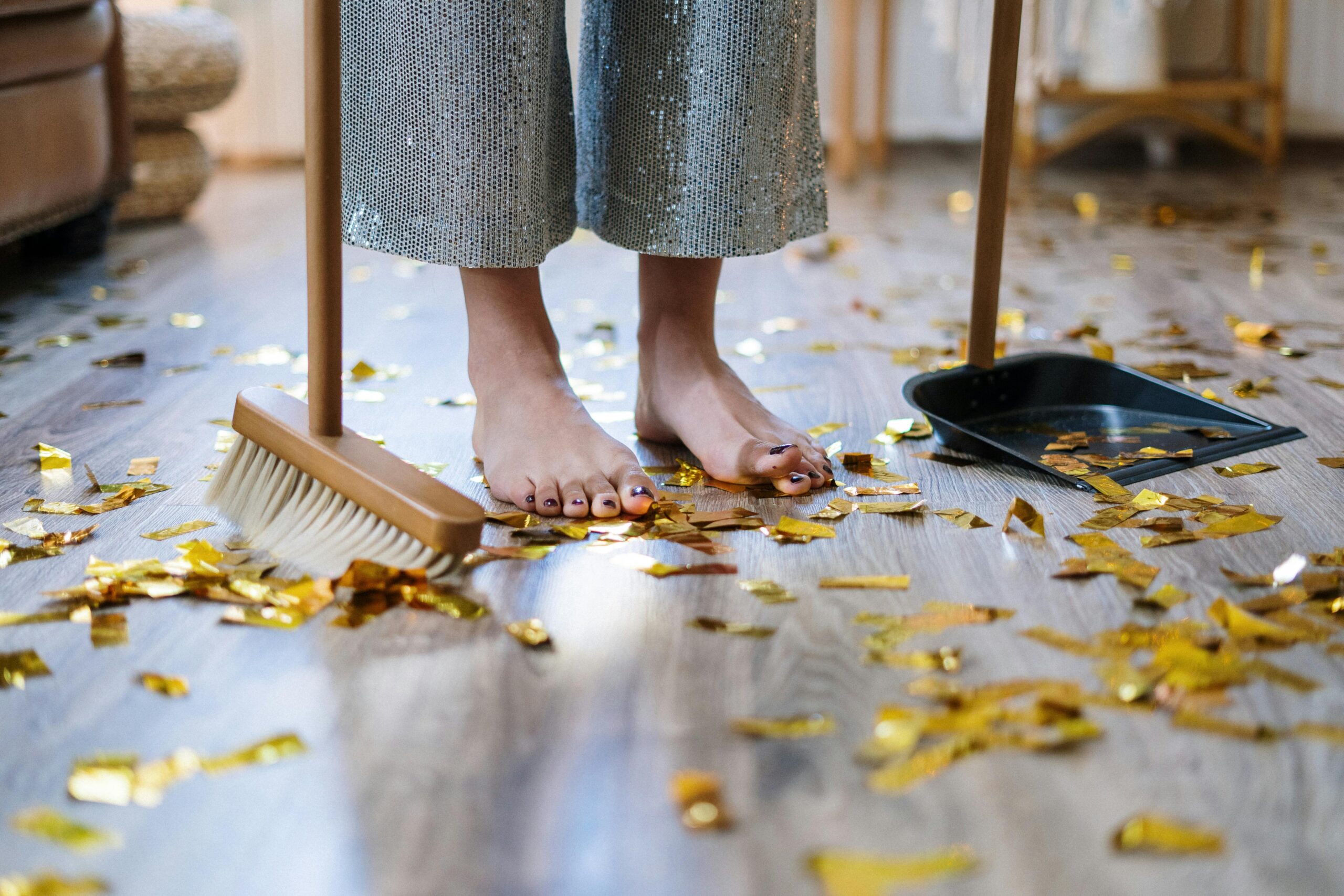 Barefoot woman sweeping gold confetti with broom and dustpan indoors.